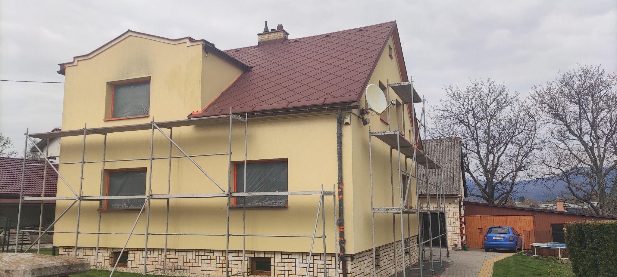 Żółty dom jednorodzinny w trakcie malowania elewacji, okna zabezpieczone folią, rusztowania rozstawione wzdłuż ścian, widoczna antena satelitarna, w tle budynki gospodarcze i drzewa.