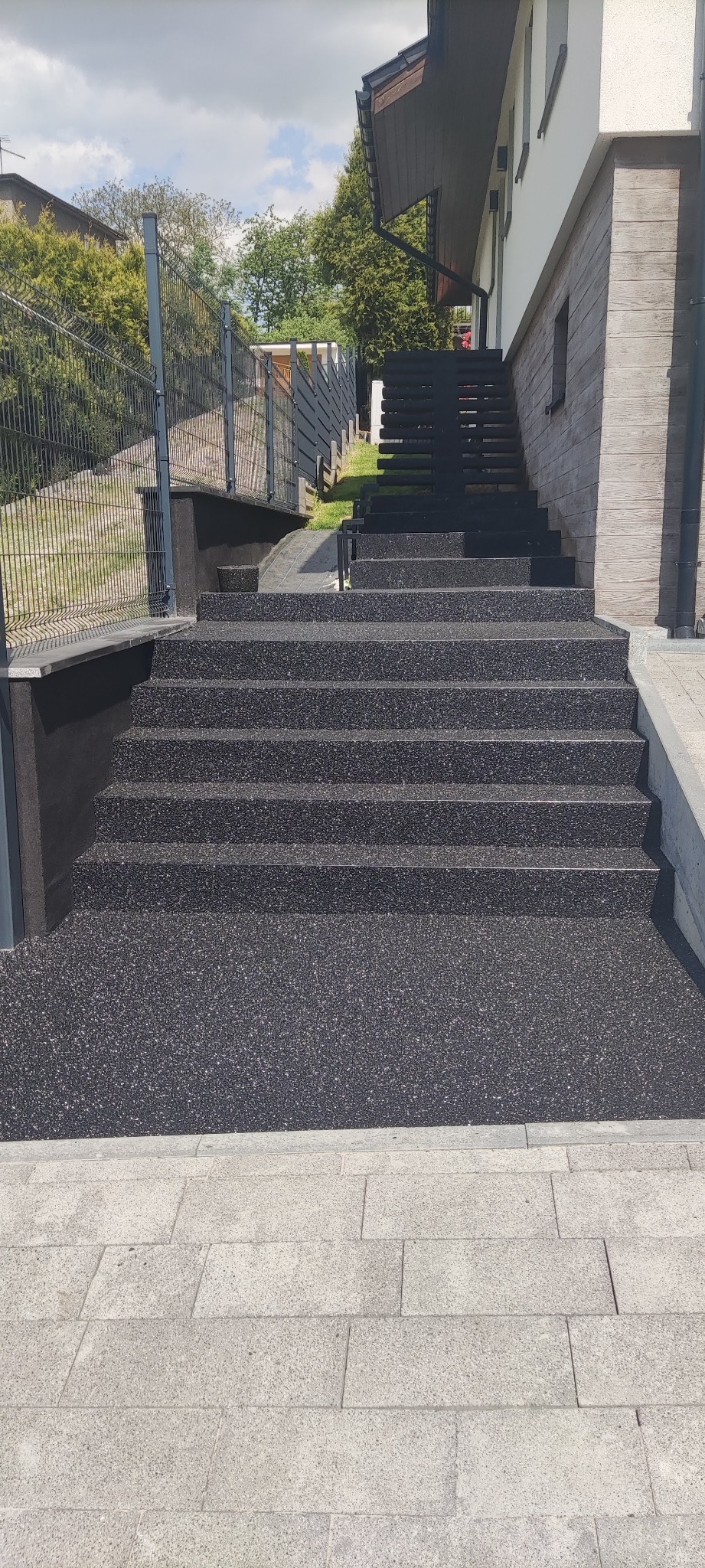 Zewnętrzne schody z czarnej, żywicznej nawierzchni prowadzące do domu z drewnianą elewacją i metalową balustradą.