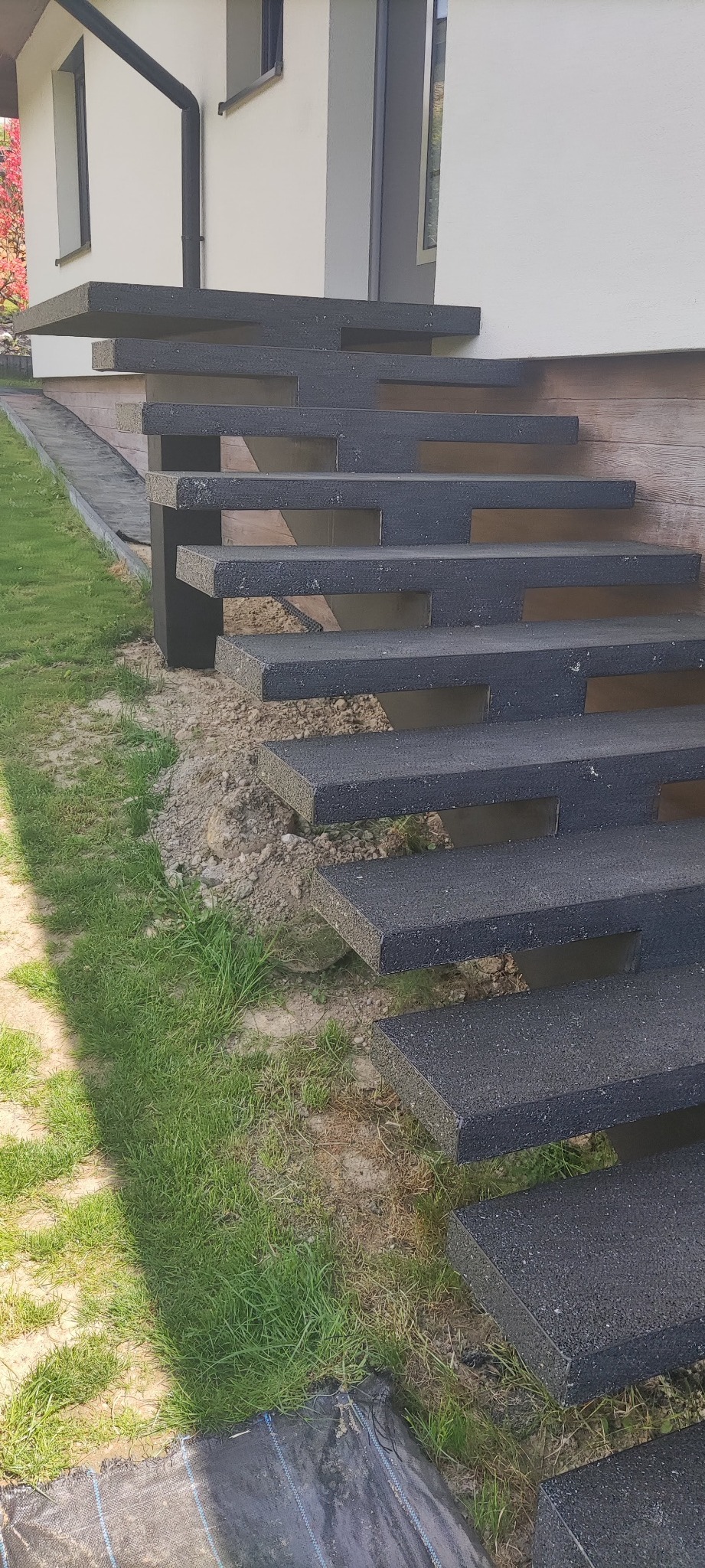 Nowoczesne schody zewnętrzne z ciemnego betonu, z geometrycznymi wycięciami pod stopniami, prowadzące do budynku z jasną elewacją i drewnianym panelem, widoczne trawnik i fragment czerwonego krzewu.