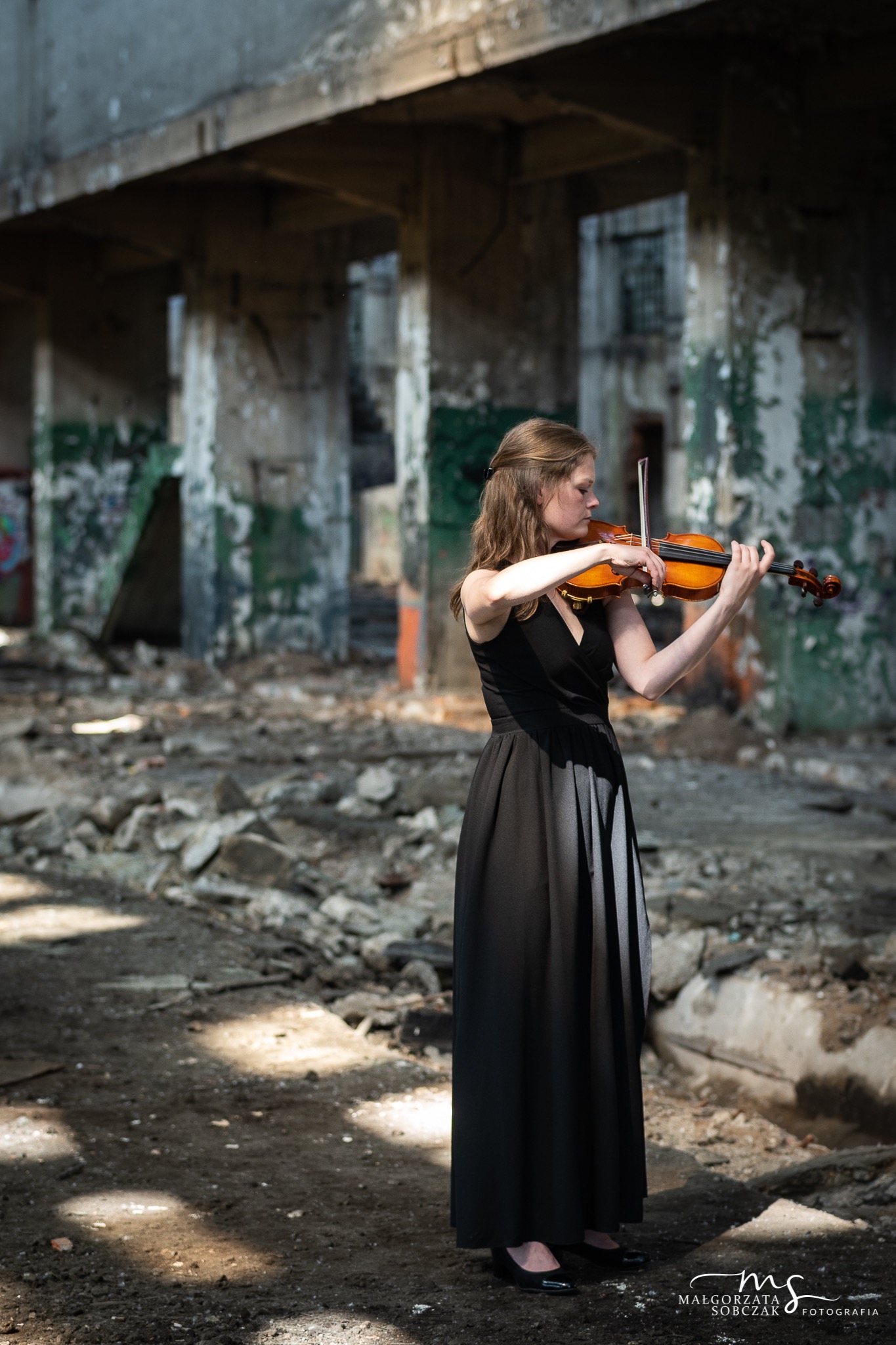 Kobieta w czarnej sukni gra na skrzypcach w opuszczonym, zrujnowanym budynku.