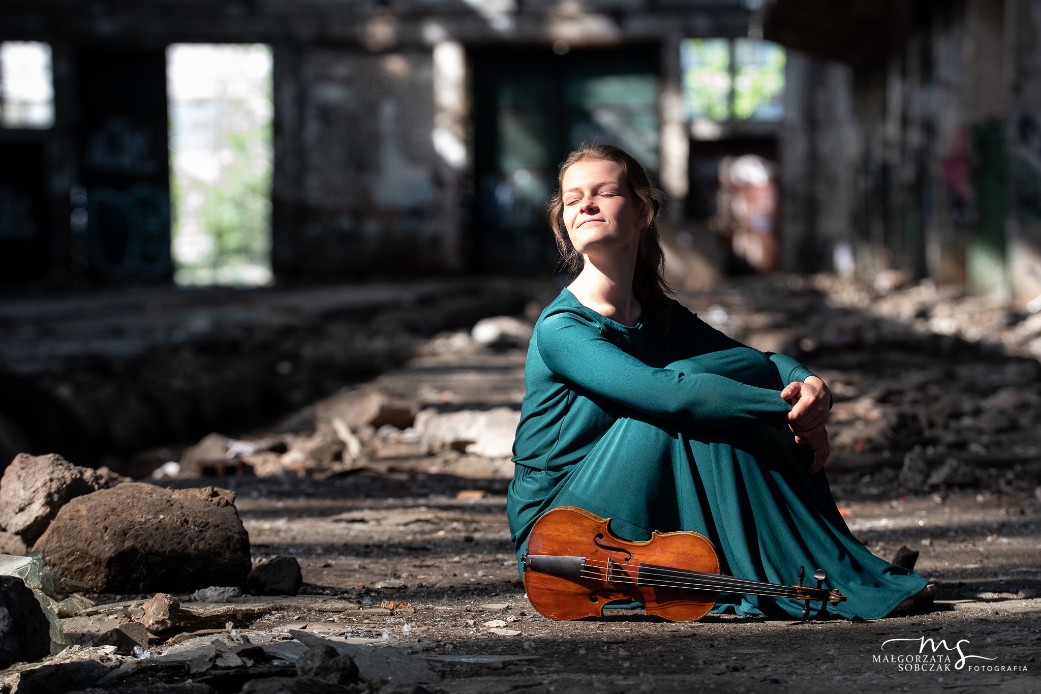 Kobieta w szmaragdowej sukni siedzi na gruzach w opuszczonym budynku, obok niej leży skrzypce w ciepłym odcieniu brązu, twarz kobiety oświetlona słońcem, oczy zamknięte.