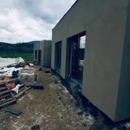 Budynek jednorodzinny w trakcie wykończenia elewacji, widoczne ocieplenie styropianem i świeżo nałożony tynk, okna z ciemnymi ramami, na ziemi materiały budowlane i palety, w tle zielone pola i las.