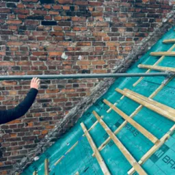 Izolacja dachu z użyciem zielonej membrany BRAAS Divoroll Universal+ i drewnianych łat, widoczne fragmenty ceglanego komina i ręka pracownika na tle nieba.