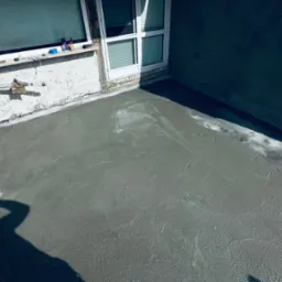 Wyrównana, świeżo wylana wylewka betonowa na balkonie, widoczne ślady po narzędziach, cień osoby fotografującej, okno balkonowe i fragment elewacji budynku w tle.