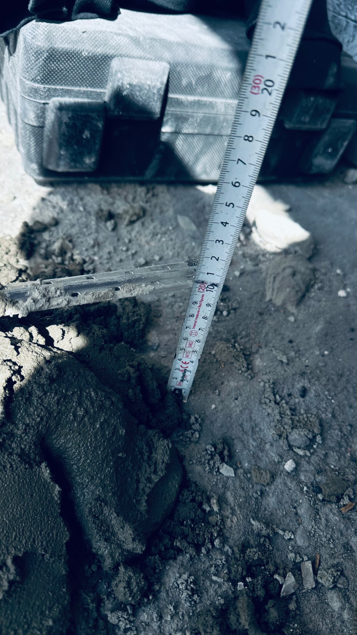 Pomiar grubości świeżo nałożonej warstwy zaprawy murarskiej za pomocą rozwijanej miarki, na tle szarej podłogi i czarnej skrzynki narzędziowej.