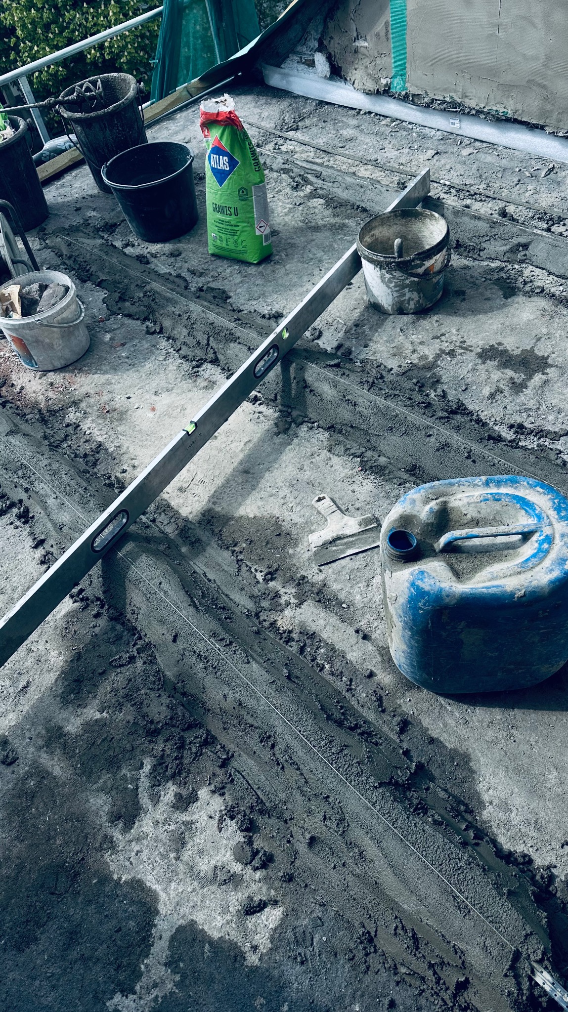 Przygotowanie podłoża pod wylewkę: widoczne wytyczone pasy zaprawy, poziomica, wiadro, paca, worek z zaprawą Atlas i kanister z wodą.