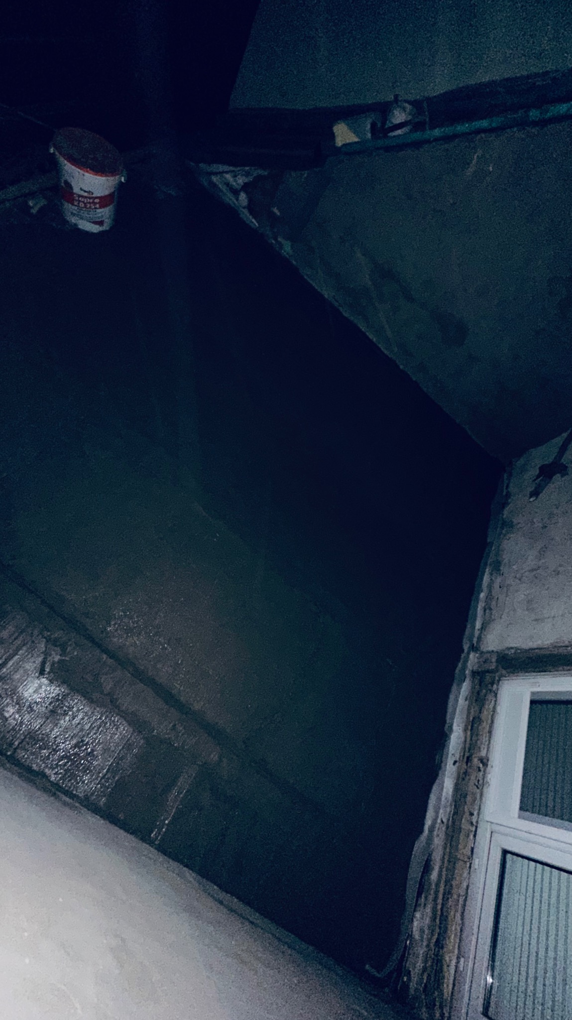 Niedokończone wnętrze z widocznym wiadrem z farbą na górze, ciemne ściany i fragment okna w rogu.