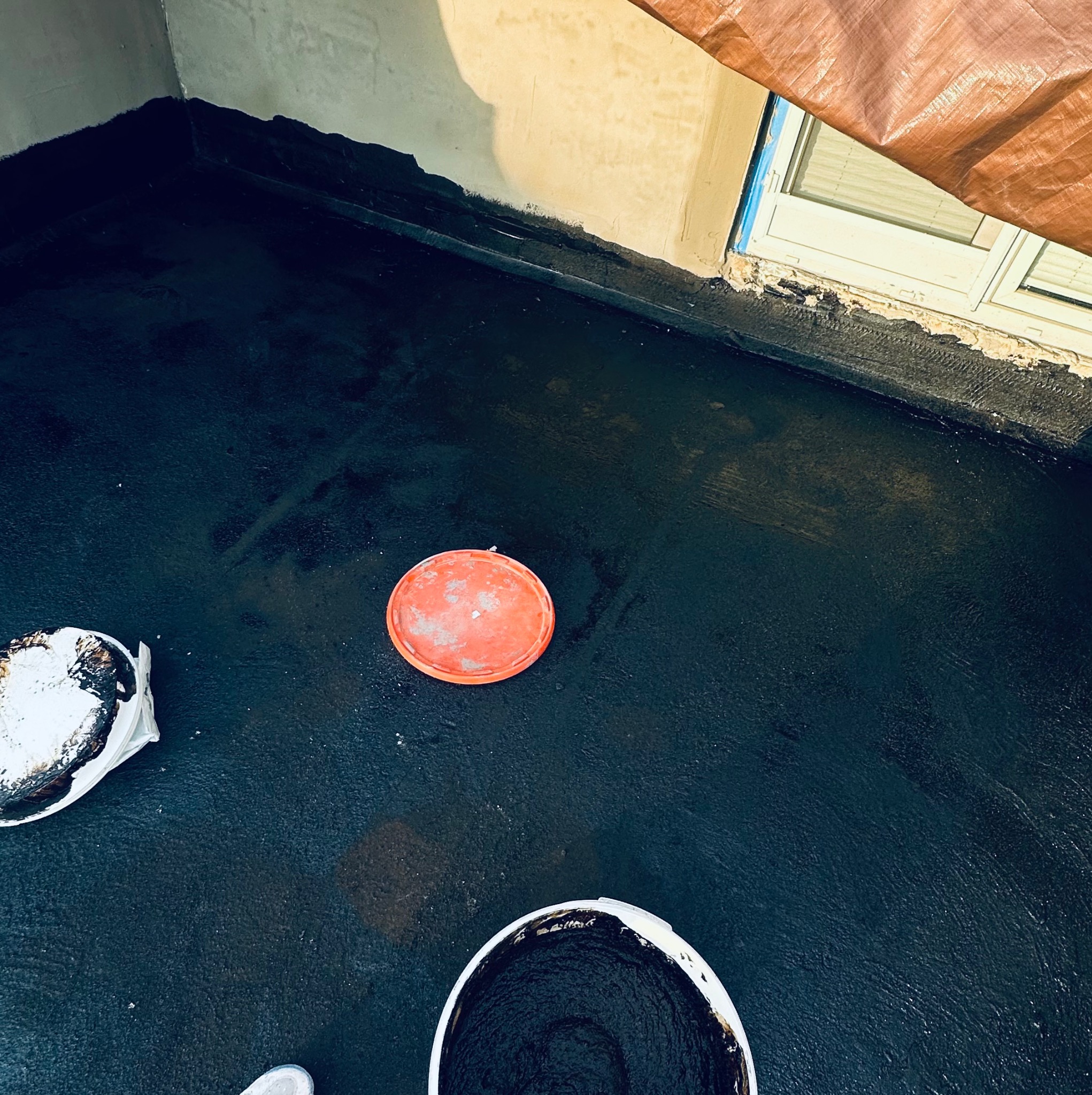 Hydroizolacja balkonu w trakcie prac: czarna powłoka, wiadro z masą bitumiczną, pomarańczowa pokrywka i zabezpieczone okno folią malarską.