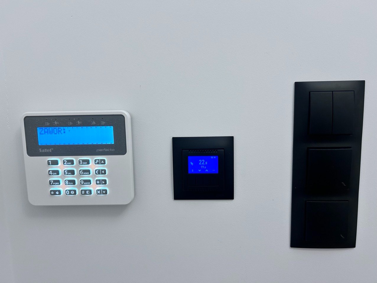 Centrala alarmowa Satel z wyświetlaczem wskazującym status zaworu, obok panel sterowania inteligentnego domu i panel włączników światła w czarnej ramce, zamontowane na białej ścianie.