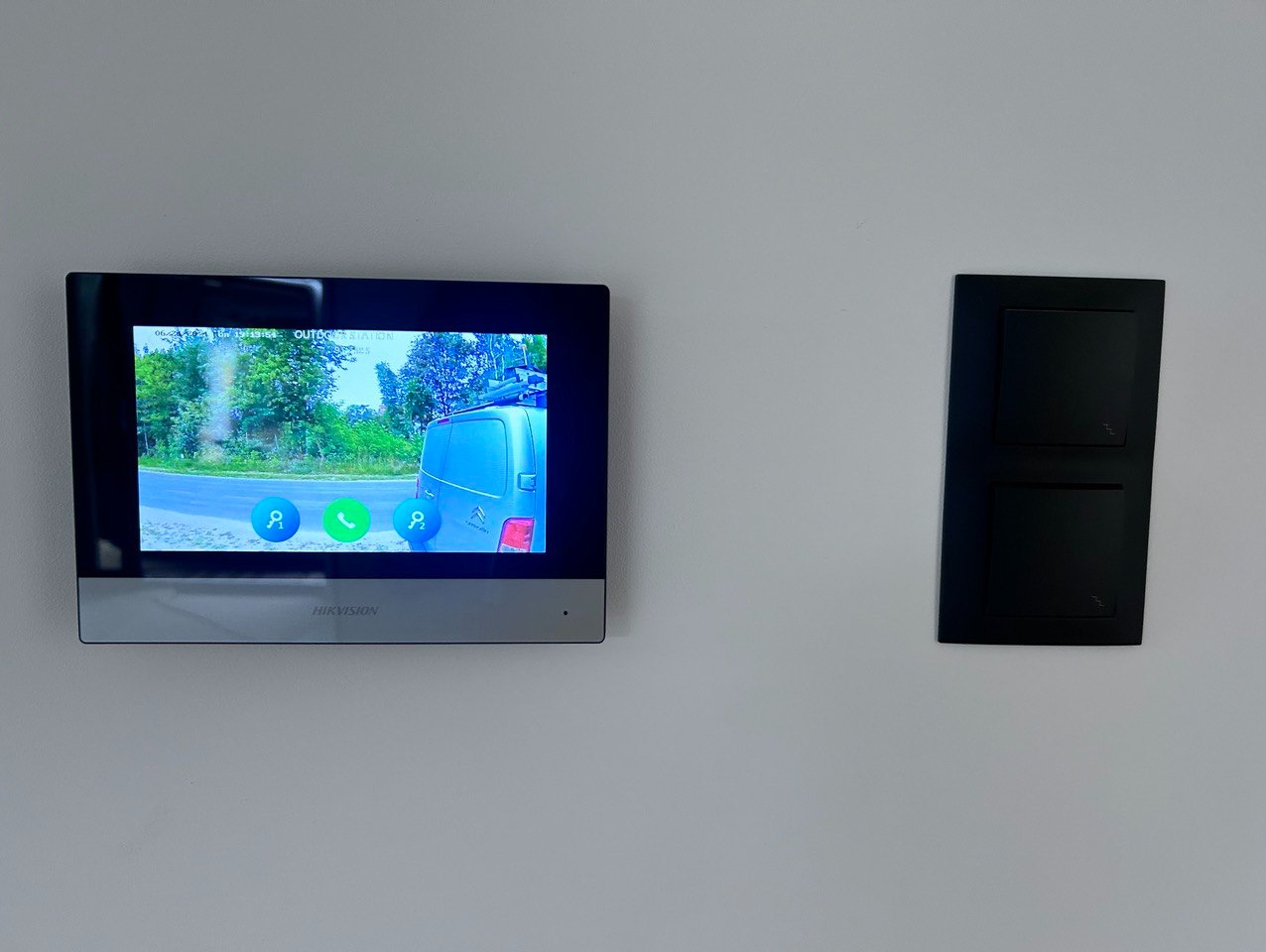Zamontowany wideodomofon IP marki Hikvision z widokiem na podjazd, gdzie stoi niebieski bus. Obok widoczny czarny włącznik światła.