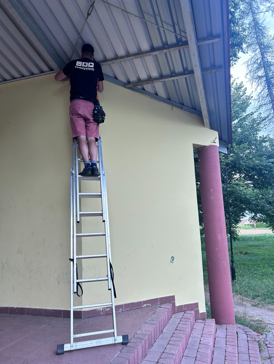 Elektryk na drabinie montuje przewody pod zadaszeniem budynku, widoczny pas z narzędziami i koszulka z logo firmy.
