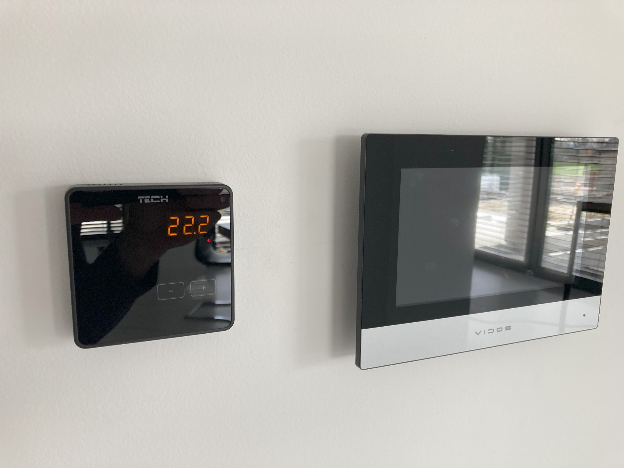 Czarny panel sterowania ogrzewaniem TECH z wyświetlaną temperaturą 22.2 stopnie Celsjusza, zamontowany obok wideodomofonu Vidos na białej ścianie, z odbiciem okna w ekranach urządzeń.