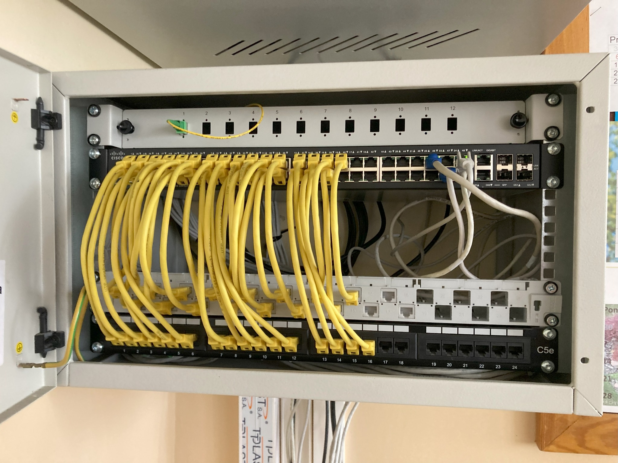 Szafa rack z panelem krosowniczym, switchem Cisco i uporządkowanymi żółtymi kablami Ethernet, widoczne oznaczenia portów i przewód uziemiający.