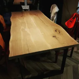 Stół z blatem z jasnego drewna o nieregularnych krawędziach, osadzony na czarnych, metalowych nogach; widoczne sęki i naturalne usłojenie drewna, na podłodze parkiet ułożony w jodełkę.