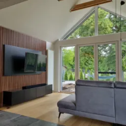 Nowoczesny salon z ceglaną ścianą, drewnianymi lamelami, wiszącym telewizorem i czarną szafką RTV, widok na ogród przez duże okna.