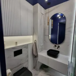 Nowoczesna łazienka z szafkami w zabudowie, umywalką nablatową, owalnym lustrem i marmurowymi płytkami na ścianach i podłodze. Granatowy sufit z designerskim oświetleniem.