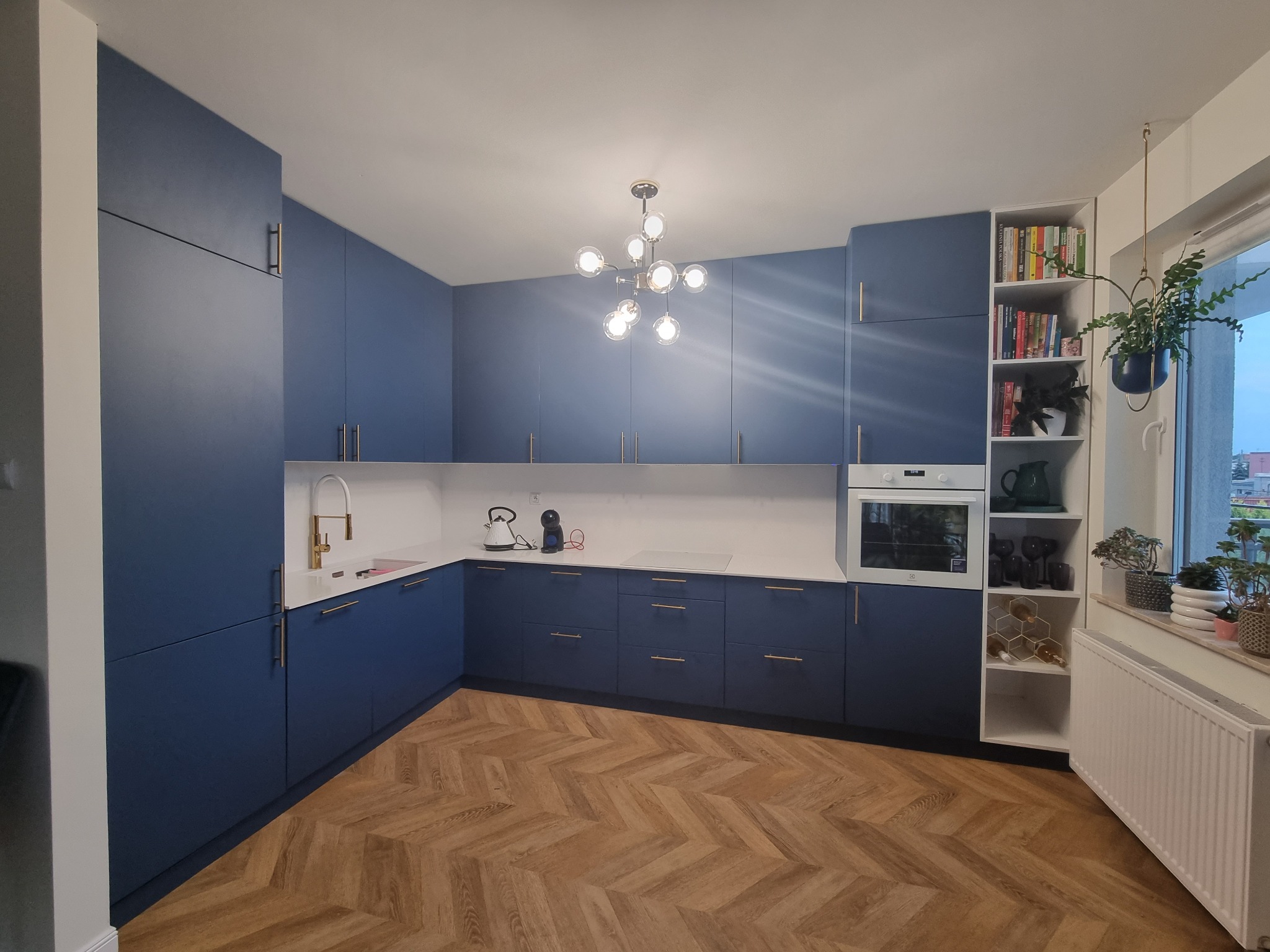 Nowoczesna kuchnia w odcieniach granatu z drewnianą podłogą w jodełkę, oświetlona designerską lampą sufitową, z białym blatem i zabudową sięgającą sufitu, uzupełniona o otwarty regał z książkami...