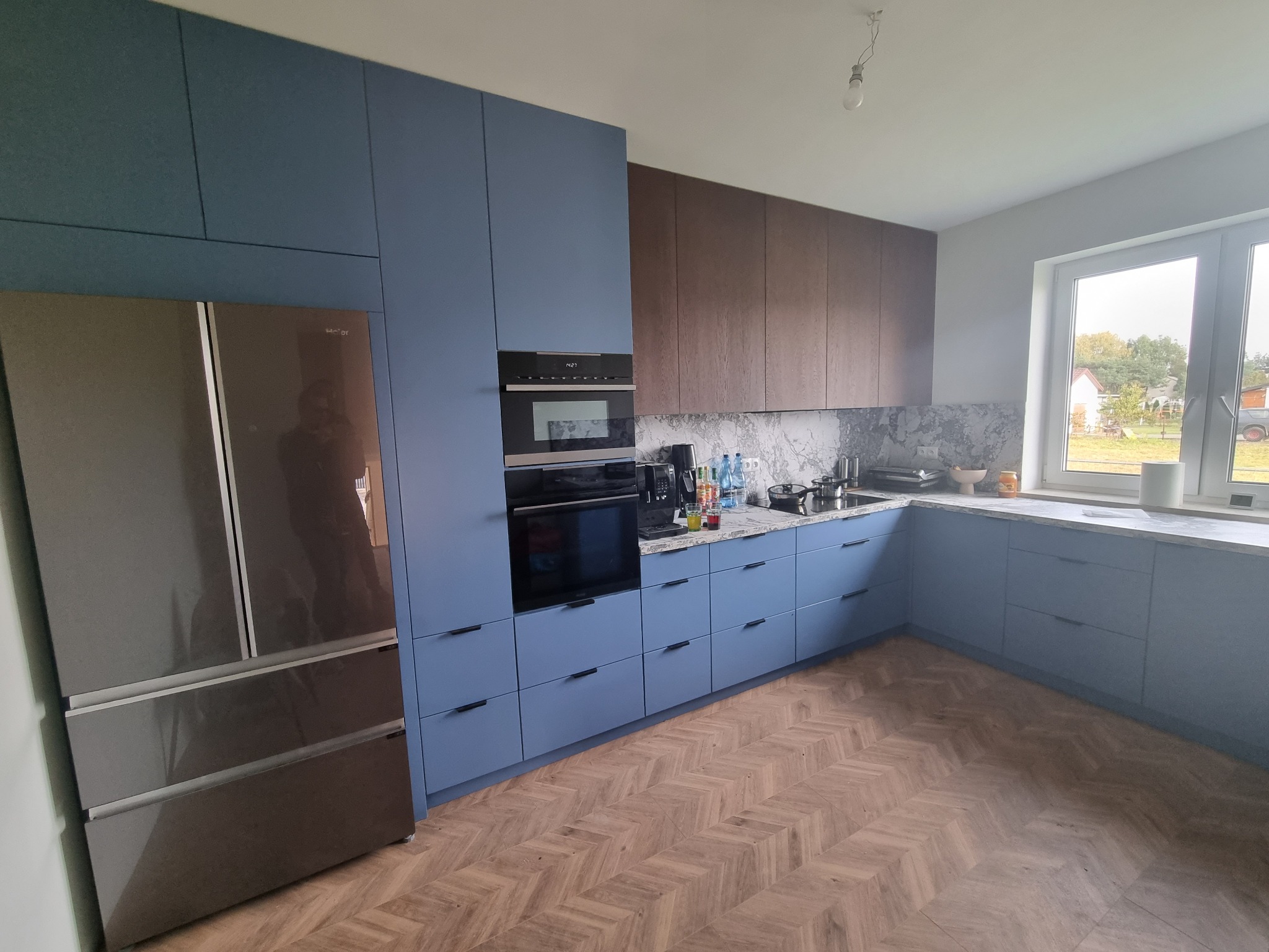 Nowoczesna kuchnia z zabudową w odcieniach niebieskiego i brązu, z parkietem ułożonym we wzór jodełki, oraz dużym, ciemnym lodówko-zamrażarką.