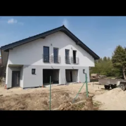 Nowo otynkowany dom jednorodzinny z szarą elewacją, czarnymi oknami i balkonami, widoczny na tle nieba i drzew, z placem budowy przed budynkiem.