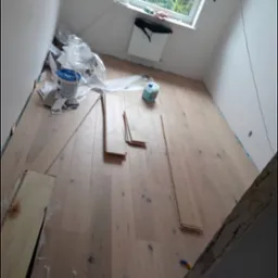 Poddasze w trakcie montażu jasnych paneli podłogowych, widoczny klej, puszka z farbą i okno dachowe.