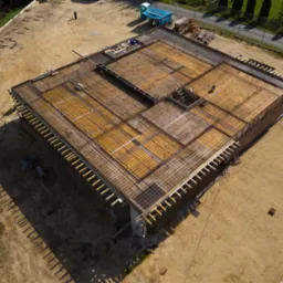 Widok z góry na plac budowy z przygotowanym szalunkiem i zbrojeniem pod fundamenty domu, widoczne deski, siatka zbrojeniowa, teren wokół wyrównany, w tle drzewa i droga.