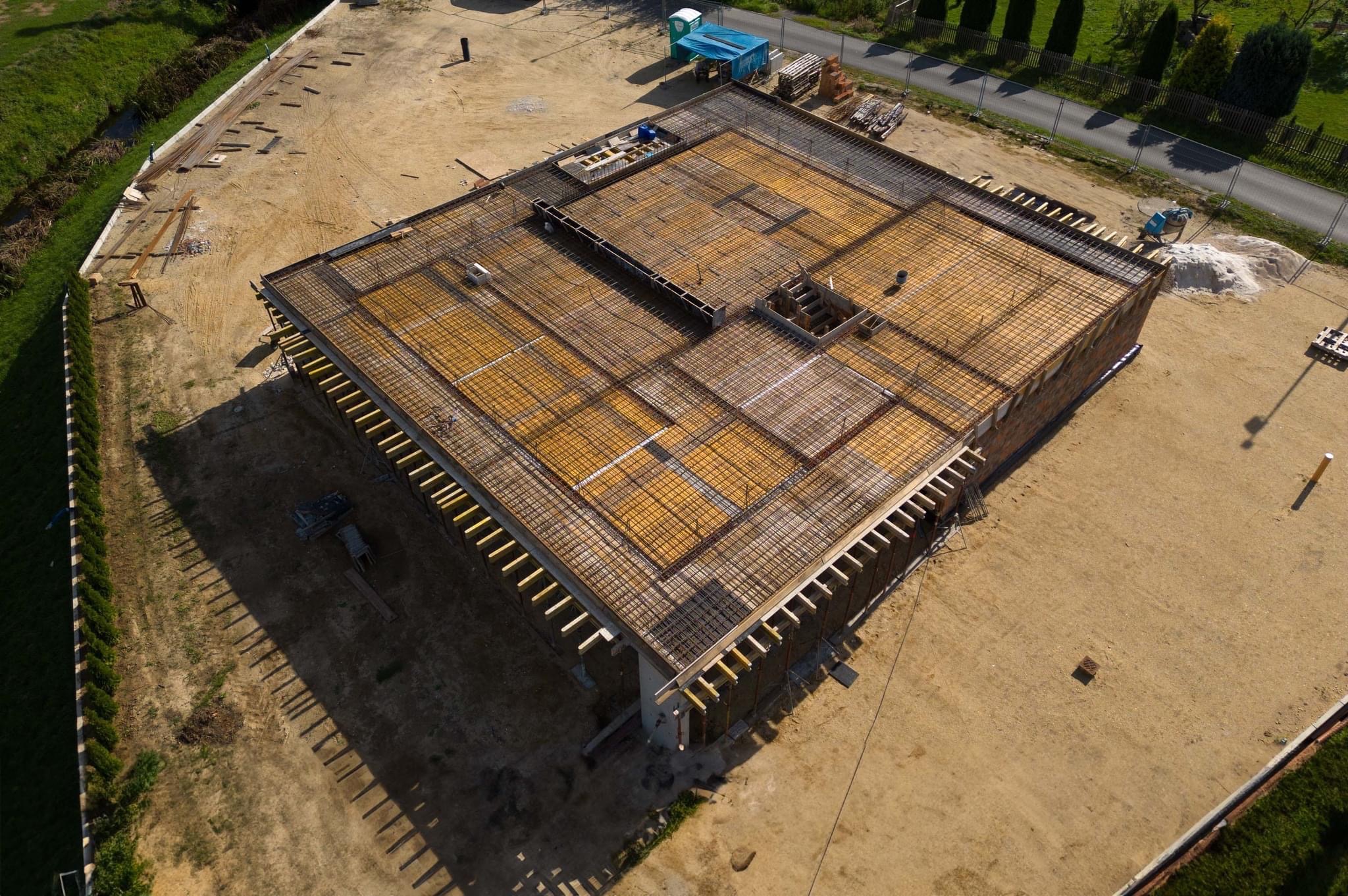 Widok z góry na plac budowy z przygotowanym szalunkiem i zbrojeniem pod fundamenty domu, widoczne deski, siatka zbrojeniowa, teren wokół wyrównany, w tle drzewa i droga.