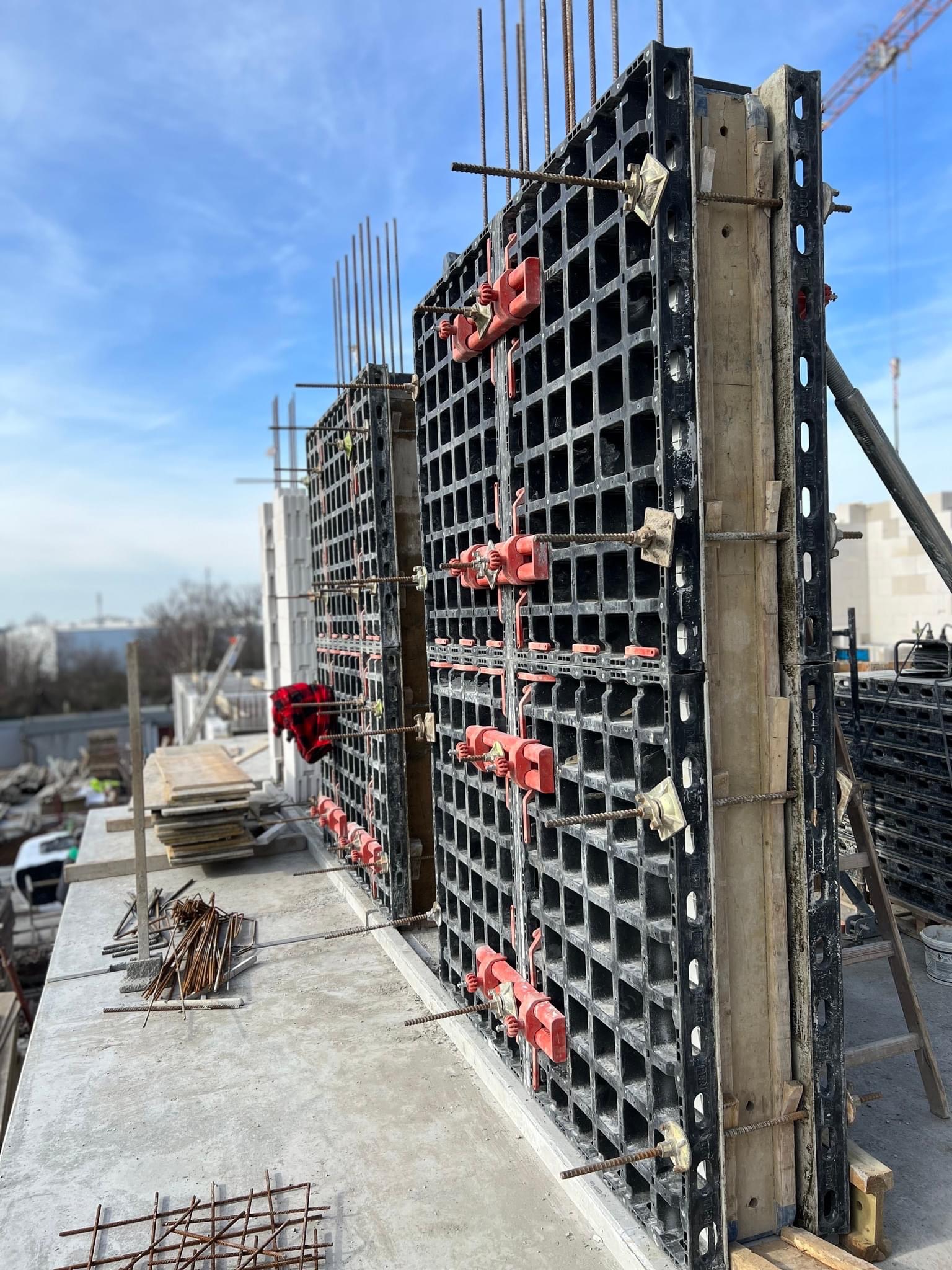 Szczegółowe ujęcie szalunków systemowych z czerwonymi zamkami, przygotowanych do zalewania betonem na budowie, widoczne wystające pręty zbrojeniowe i elementy konstrukcji w tle.