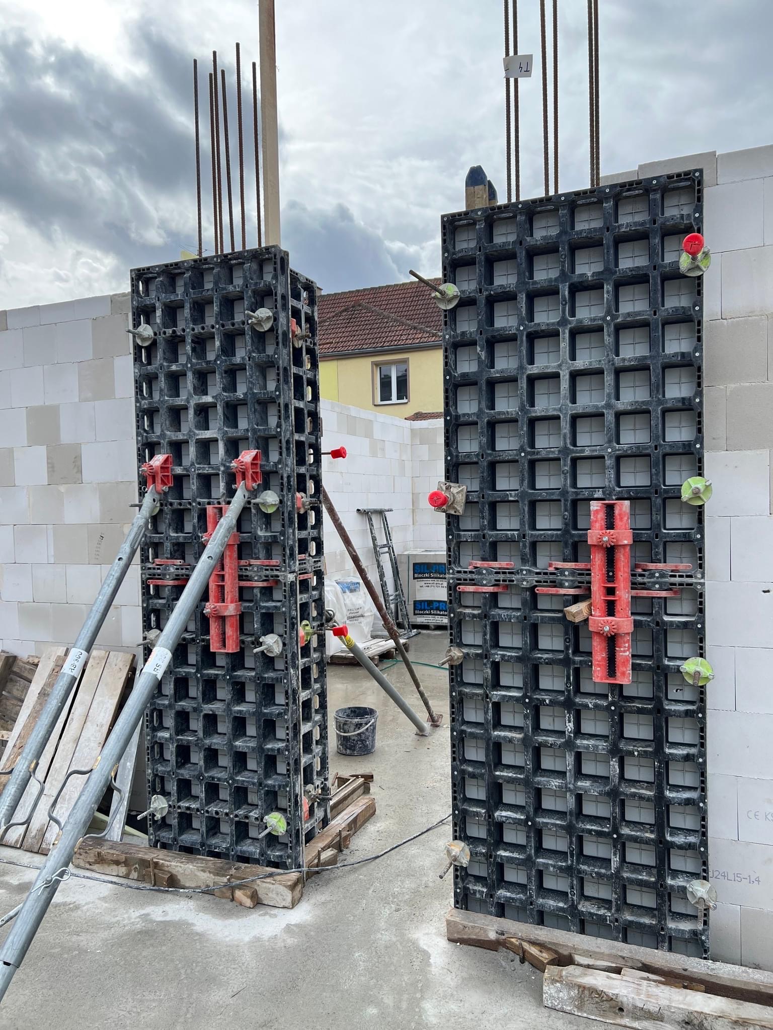 Szare, modułowe szalunki kolumnowe, podparte metalowymi stemplami, z wystającymi prętami zbrojeniowymi, ustawione przy ścianie z białych bloczków betonu komórkowego na budowie.