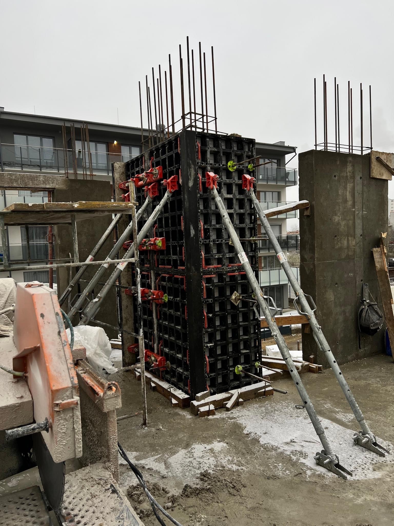 Szare, pochmurne niebo nad budową: szalunek kolumny żelbetowej z wystającymi prętami zbrojeniowymi, wsparty na metalowych podporach, obok widoczny fragment obudowy kolumny po zalaniu betonem...