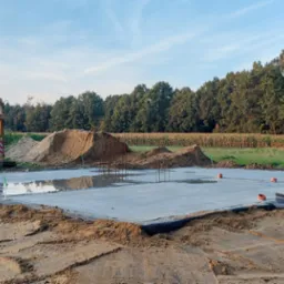 Wylany fundament pod dom jednorodzinny, z widocznym zbrojeniem, częściowo zalany wodą, z koparko-ładowarką w tle, na obrzeżach pola kukurydzy.