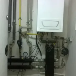 Biały piec grzewczy z podłączeniami rur, zaworów i przewodów, widoczne rury odprowadzające spaliny, na podłodze zielone wiadro z wodą i wąż, obok biała umywalka z ręcznikiem.