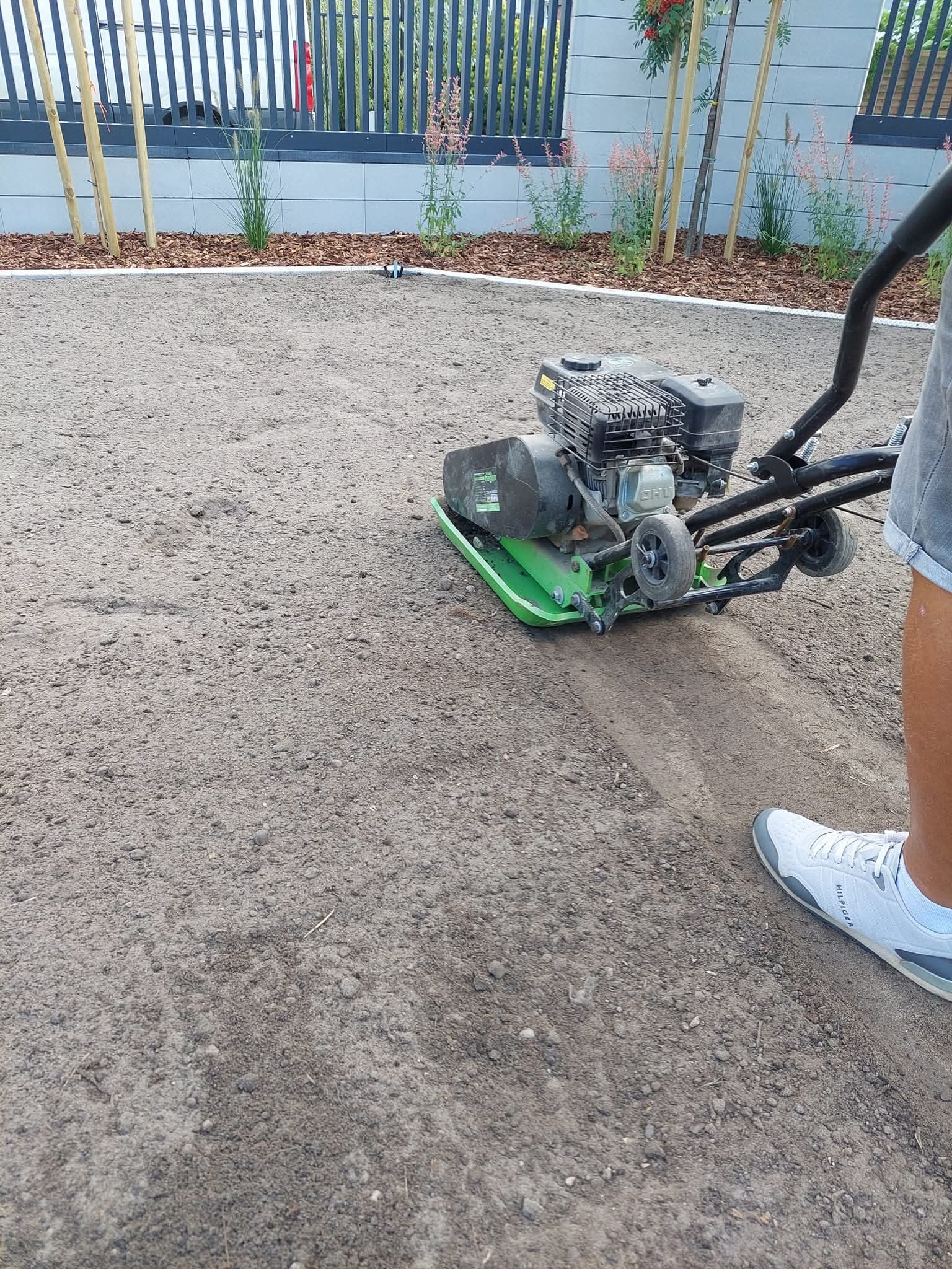 Praca zagęszczarką na przygotowanym podłożu pod trawnik, widoczny fragment buta osoby obsługującej maszynę oraz rabata z korą i roślinami w tle.