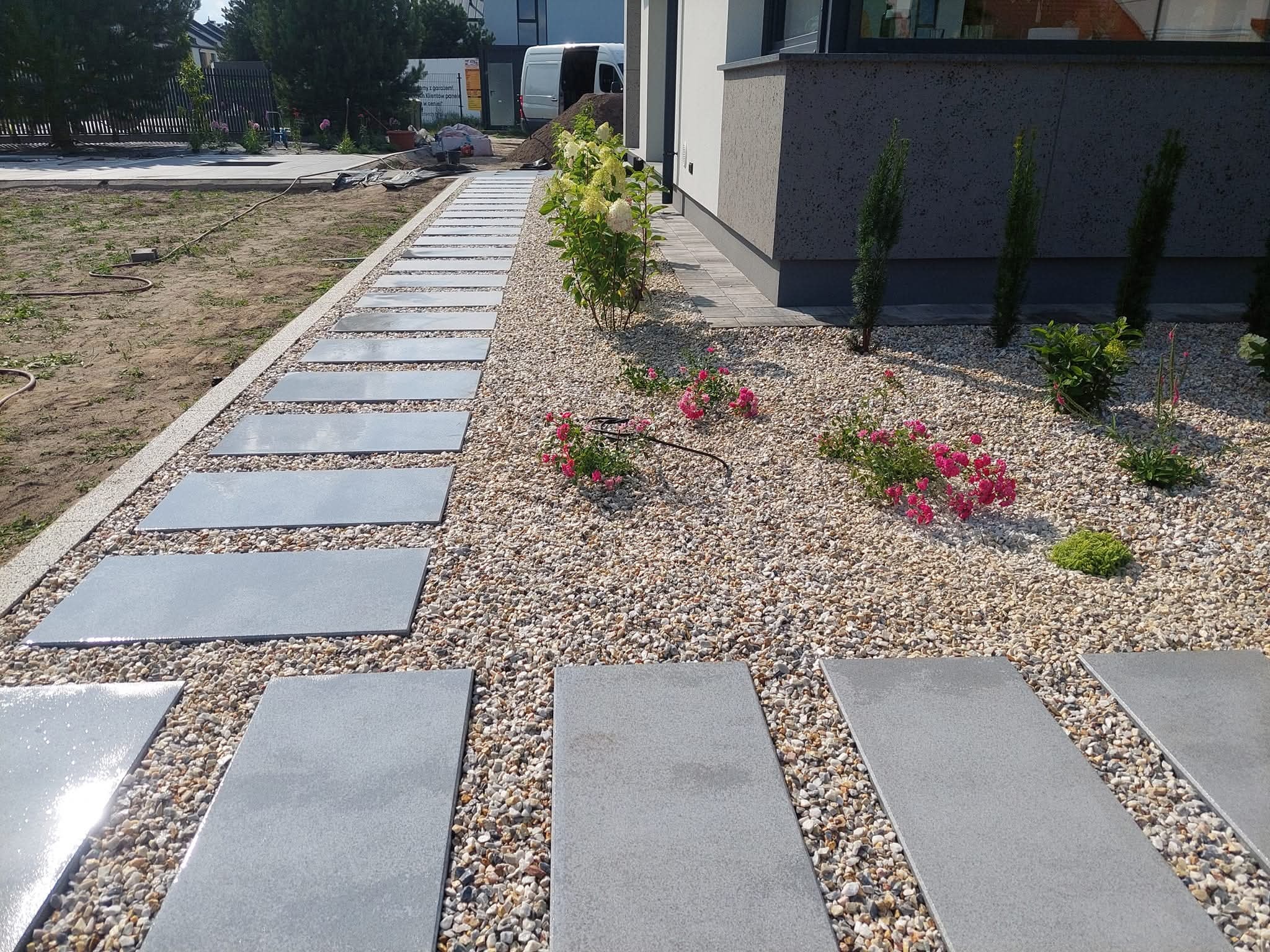 Ścieżka z płyt betonowych w żwirowym ogrodzie, obok domu z nasadzeniami roślin. Nowoczesny design z elementami zieleni i kamienia.