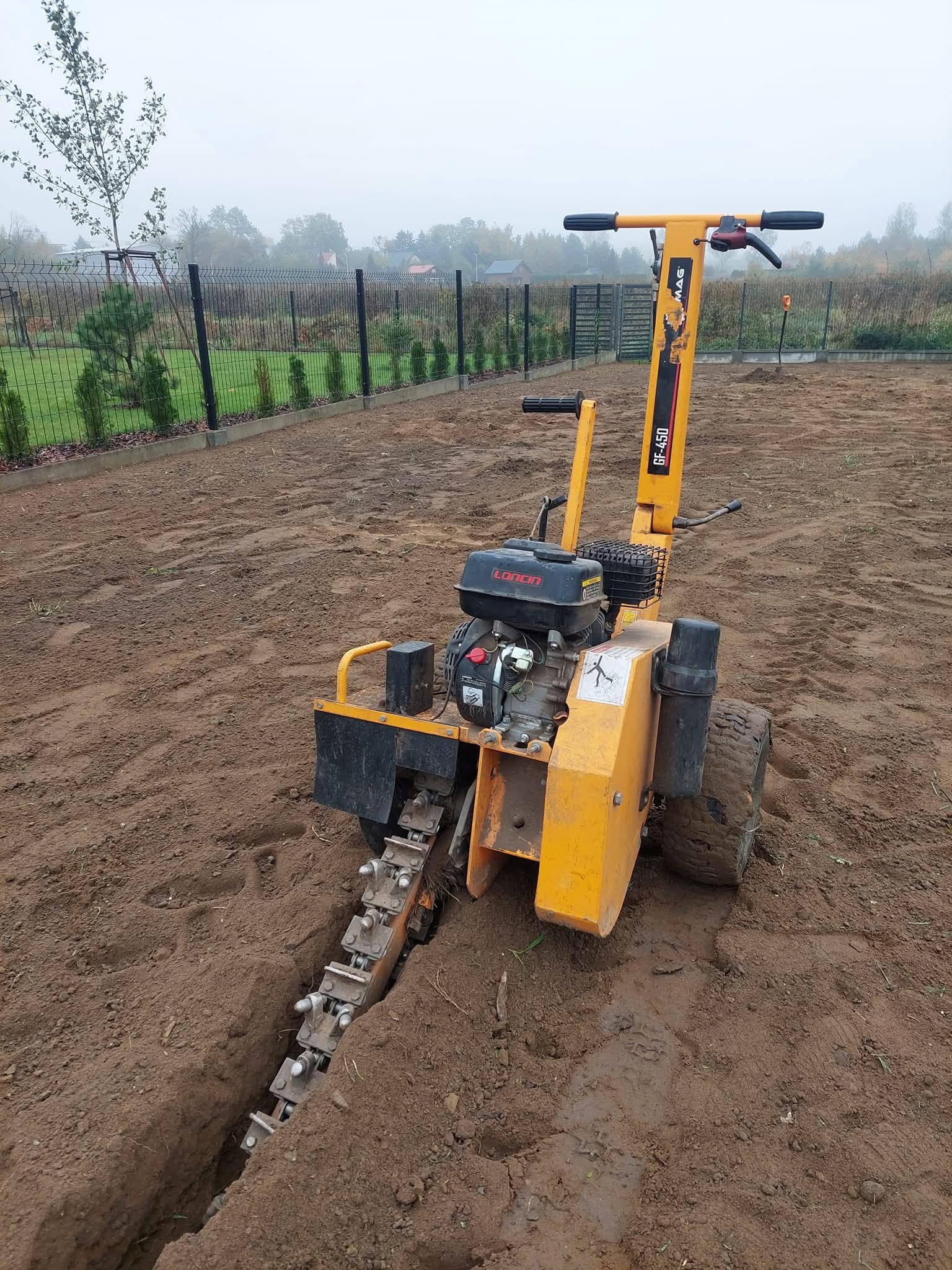 Żółta glebogryzarka MAG GF-450 w trakcie pracy, tworząca wąski wykop w przygotowanej ziemi pod system nawadniania lub instalację, z widocznym ogrodzeniem w tle.