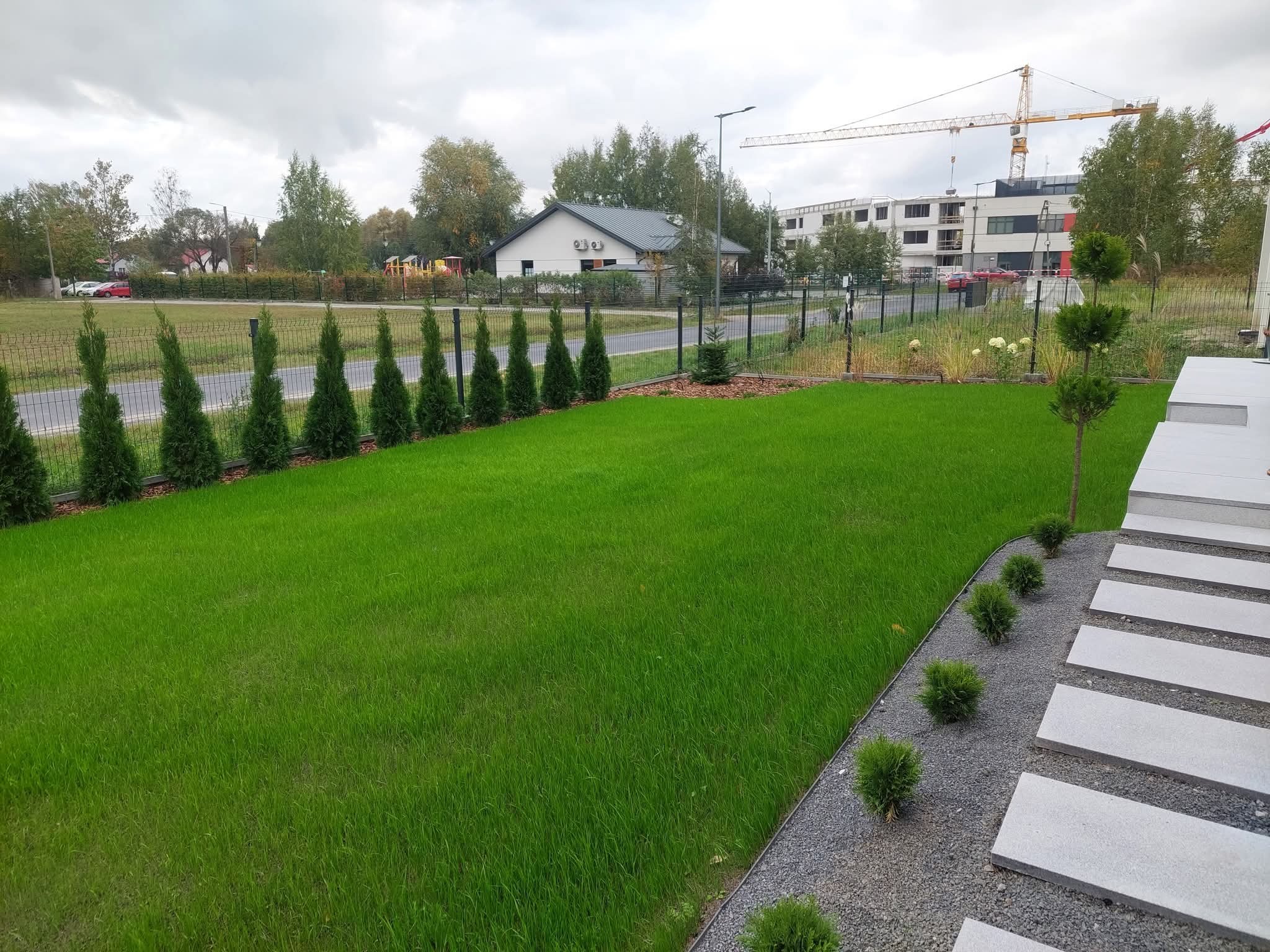 Zadbany trawnik z równo przyciętą trawą, obrzeżem z kamyków i ścieżką z płyt. W tle rząd tuj i budynek mieszkalny z placem zabaw. Widoczny żuraw budowlany.