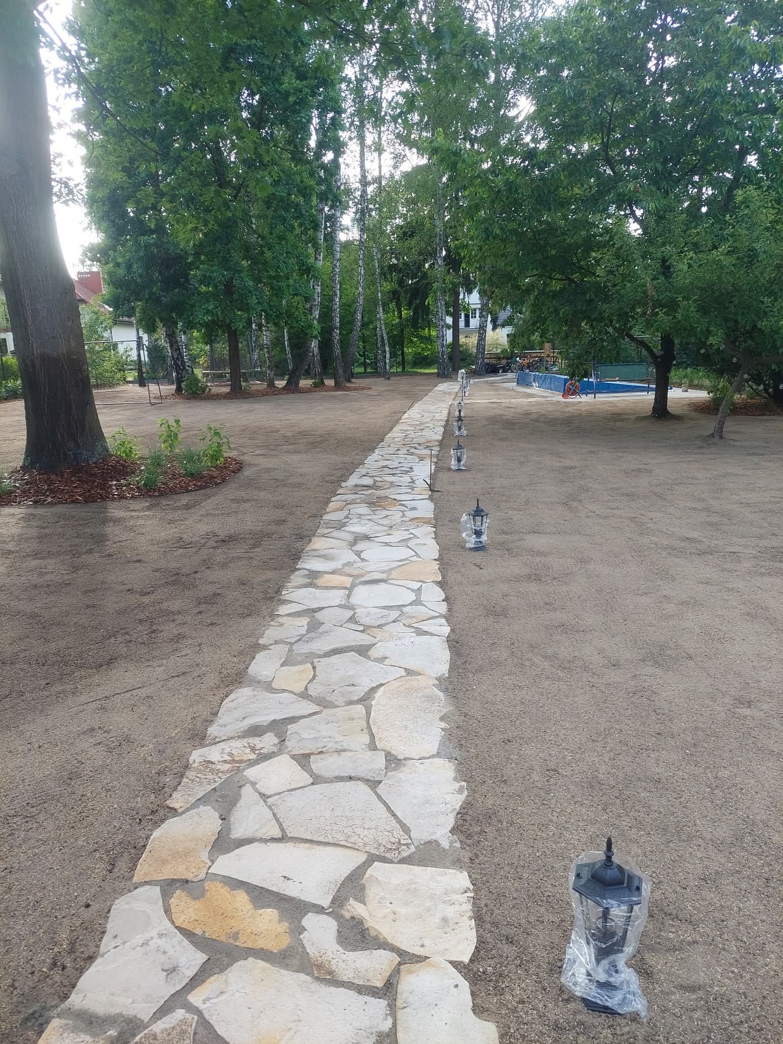 Ścieżka z nieregularnych płyt kamiennych prowadząca przez przygotowany teren z młodymi drzewami i lampami ogrodowymi w folii. W tle basen i budynki.