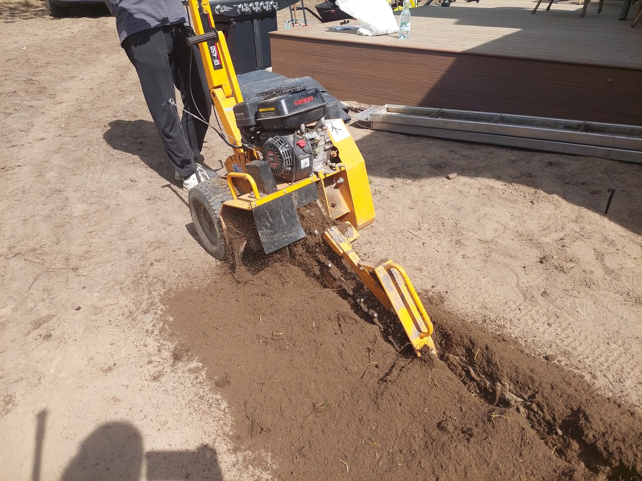 Żółta glebogryzarka GF-450 w trakcie pracy, tworząca wąski rów w ziemi. Operator w sportowych butach i dresach obok maszyny. W tle grill i fragment drewnianego podestu.