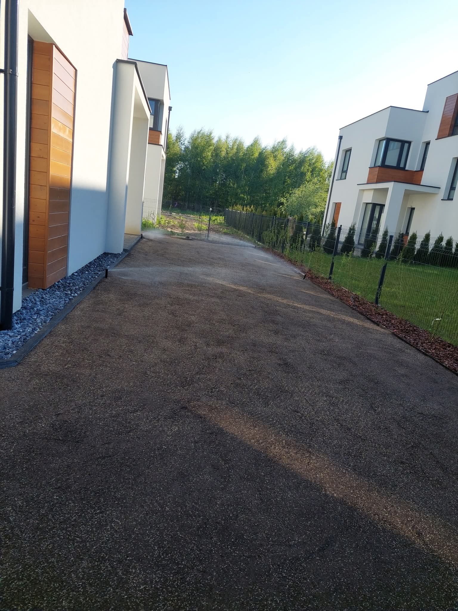 Świeżo zasiana trawa na posesji między dwoma nowoczesnymi domami, z systemem nawadniania w trakcie pracy. Widok na trawnik i ogrodzenie.