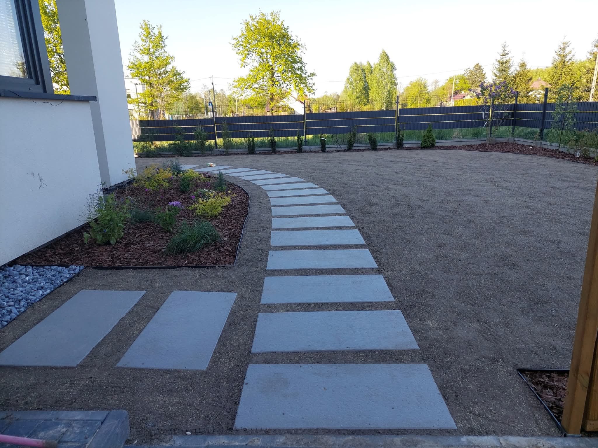 Ścieżka z płyt prowadząca przez świeżo założony trawnik, obok rabaty z korą i roślinami, w tle ogrodzenie i drzewa. Widok na ogród.