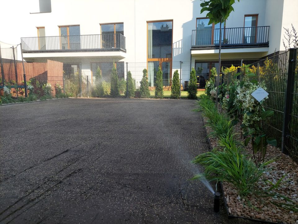 Automatyczny system nawadniania w trakcie pracy na przygotowanym podłożu pod trawnik, widoczne zraszacze i nawilżona gleba, w tle nowoczesny budynek z balkonami.