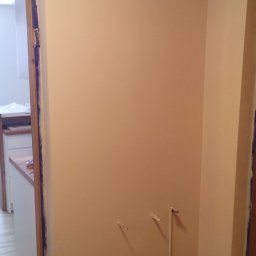 Usługi Remontowo Budowlane Przemysław Węgrzyn - Świeżo pomalowane ściany w odcieniach beżu w trakcie prac wykończeniowych, zabezpieczone folią i taśmą malarską, widoczne wystające rury instalacji wodnej i szafka z kluczem.