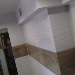 Usługi Remontowo Budowlane Przemysław Węgrzyn - Wykończona łazienka z białymi i brązowymi płytkami imitującymi drewno, widoczny fragment lustra z odbiciem osoby robiącej zdjęcie telefonem.