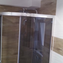 Usługi Remontowo Budowlane Przemysław Węgrzyn - Kabina prysznicowa z przesuwnymi drzwiami, wykończona płytkami imitującymi drewno i białymi płytkami z delikatnym wzorem, oświetlona górnym światłem.