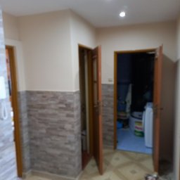 Usługi Remontowo Budowlane Przemysław Węgrzyn - Korytarz po remoncie z jasnymi ścianami, płytkami imitującymi kamień na dole i wzorzystą podłogą. Otwarta drewniana futryna drzwi ukazuje wnętrze łazienki z pralką i pojemnikami z detergentami.