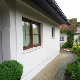 Boczny widok odnowionego domu z białą elewacją i brązowymi oknami, z widocznym fragmentem chodnika z kostki brukowej i zielenią ogrodu.