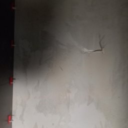 Usługi Remontowo Budowlane Przemysław Węgrzyn - Ściana w trakcie prac wykończeniowych w Jaśle: widoczne świeżo nałożone warstwy gładzi i czerwone klipsy do poziomowania płytek na krawędzi.