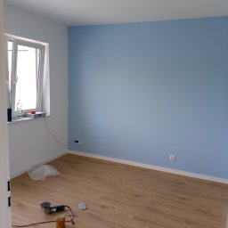 Usługi Remontowo Budowlane Przemysław Węgrzyn - Pokój po malowaniu z jedną ścianą w kolorze błękitnym, widoczne narzędzia malarskie na podłodze z paneli, okno z butelką wody na parapecie.