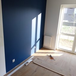 Usługi Remontowo Budowlane Przemysław Węgrzyn - Świeżo pomalowana na granatowo ściana w jasnym pokoju z drewnianą podłogą w trakcie montażu, widoczne listwy przypodłogowe i tuba z klejem.