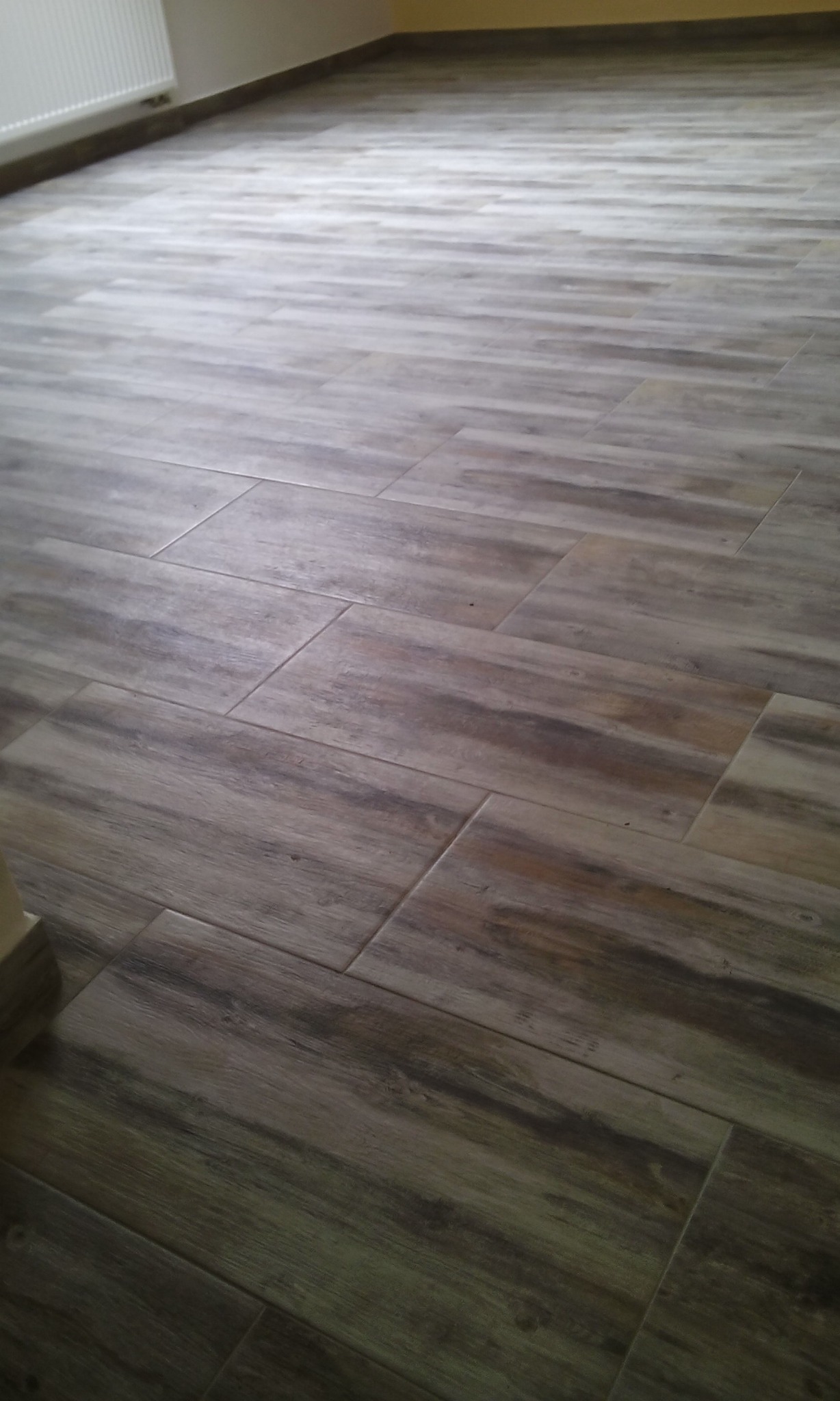 Ułożona podłoga z brązowych płytek imitujących drewno, widoczny fragment grzejnika i ściany w tle.