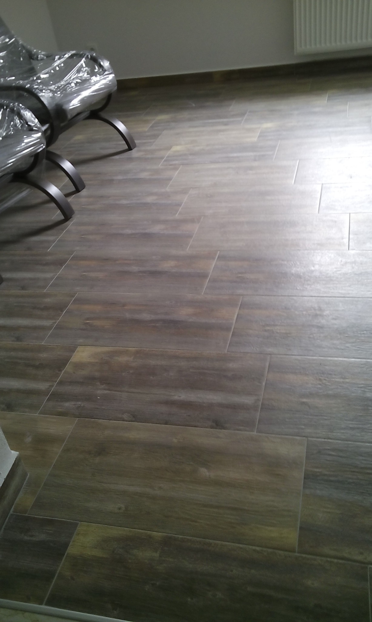 Ułożona podłoga z płytek imitujących drewno w odcieniach brązu i szarości, z widocznymi krzesłami przykrytymi folią ochronną i grzejnikiem.